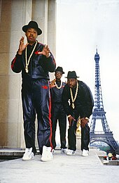 Three men wearing tracksuits, bowler hats, and gold chains looking into the camera. The Eiffel Tower is in the background.