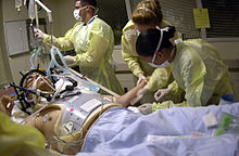 Health care providers attending to a person on a stretcher with a gunshot wound to the head; the patient is intubated, and a mechanical ventilator is visible in the background