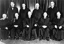 Five men sitting in a row with four men standing behind them. All wear flowing back robes and a large black curtain is behind them