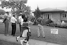 A black and white photograph of a suburban house with minor bomb damage to the roof and two windows while five black Birmingham residents stare at the damage; the yard is cordoned off with a sign saying "Danger Keep Out"