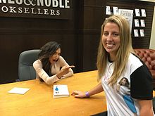 A young dark-haired woman sits at a desk in a bookstore, pointing to a blonde woman wearing her likeness on a t-shirt.
