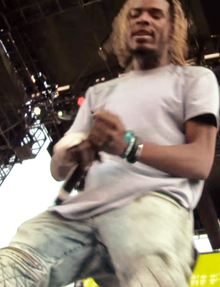 Slightly blurry image of a man on stage with a mic wearing jeans and a t-shirt.