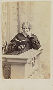 Photographie sépia d'une femme blanche d'âge mûr portant un bonnet blanc, assise et lisant sur un balcon