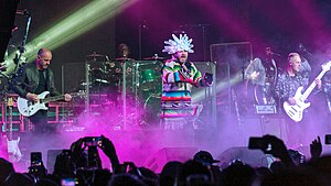 The band Jamiroquai performing on stage at Coachella 2018. At center is vocalist Jay Kay wearing a colorful Native-American shirt with frills around the waist and a white LED head-dress. Also present are a guitarist, a bassist, a drummer and two female backing vocalists. The band is engulfed in roughly waist-level stage fog colored purple by stage lighting.