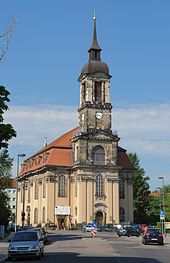 Barevná fotografie s pohledem na omítnutý barokní kostel s výraznou věží z pískovcových kvádrů s oválnou helmicí