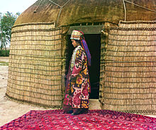 Full-length profile portrait of a woman, standing on a carpet at the entrance to a yurt, dressed in traditional clothing and jewelry
