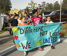 A group of students and their family members carrying a sign saying "Different Not Less: We Are United" with different coloured puzzle pieces on the sign