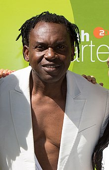 Dr. Alban squinting into camera, wearing an open jacket, partially exposing chest; two people appear to have a hand on each of his shoulders