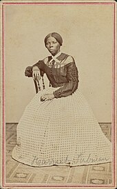 Photographie sépia en pied d'une femme noire assise sur une chaise, un coude posé sur le dossier, l'autre main sur le genou, son chemisier est sombre, sa large jupe longue est claire