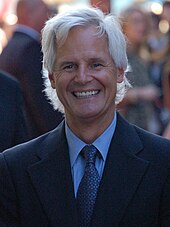 A man with long white hair and a black suit, with a tie, is standing and smiling.