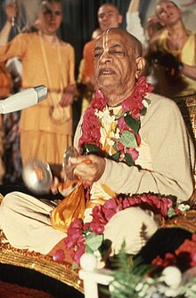 A portrait of an elderly Indian man in light-saffron robes with a red flower garland around his neck, sitting cross-legged with his eyes closed, playing hand cymbals and signing.