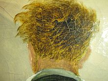 Farbige Nahaufnahme eines Hinterkopfs mit blonden, lockigen Haaren, die vom Wind verweht sind. Der Kragen vom Hemd und die Jacke sind am unteren Bildrand zu sehen. Der Hintergrund ist beige.