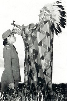 Photo of Mary Greyeyes kneeling on ground while a man in traditional Plains Cree Chief regalia waves a hand over her forehead