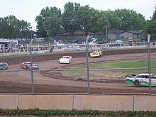 Cars race around a dirt course behind a tall metal fence.