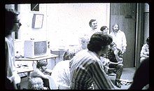 B/w photograph of a group of male people dressed in casual shirts, sitting in a circle in a room looking in the same direction. The room is furnished with desks and a TV-set.