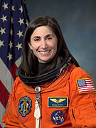 A female Caucasian astronaut wearing her orange mission suit with the American flag embroidered on her shoulder, and name tag and mission patches embroidered on her chest.