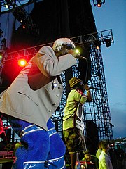 Два афро-американських чоловіки читають реп в мікрофон на сцені. Один у білявій перуці, сірій куртці та синіх штанях, а на другому зелена картата шапка, біла сорочка та шорти хакі.
