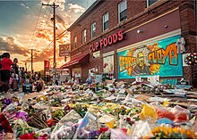 Large area of sidewalk covered in flowers and other tributes beside a building with a mural painted on the wall