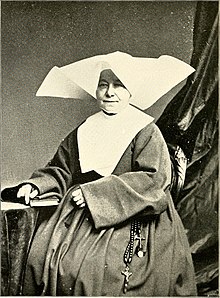Older white woman, seated, in religious habit; she has a wide white wimple on her head.