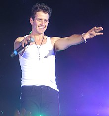 Joey McIntyre wearing a white tank top and dark pants, holding a microphone onstage, with arms outstretched toward audience
