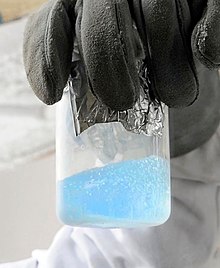 A transparent beaker containing a light blue fluid with gas bubbles.