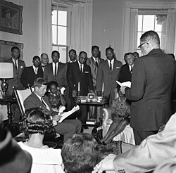 President Kennedy (seated in rocking chair); Bishop Stephen G. Spottswood, Chairman of the NAACP Board of Directors (standing right, back to camera). Standing, rear (L – R): Medgar Evers, Mississippi NAACP Field Secretary; Calvin Luper, Oklahoma City NAACP Youth Council President; Edward Turner, President of Detroit NAACP branch; Rev. W.J. Hodge; Dr. S.Y. Nixson; C.R. Darden, President of Mississippi NAACP State Conference branches; Kelly M. Alexander, member of NAACP Board of Directors; Kivie Kaplan, Chairman of NAACP Life Membership Committee; others unidentified (some partially hidden). Oval Office, White House, Washington, D.C.
