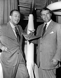 Volts Disnejs, pa kreisi, un Verners fon Brauns, pa labi. Dr. Verners fon Brauns, toreizējais armijas ballistisko raķešu aģentūras (ABMA) vadīto raķešu izstrādes operāciju nodaļas vadītājs armijas postenī Redstone Arsenal, Alabamas štatā, kuru 1954. gadā apmeklēja Volts Disnejs. 1950. gados fon Brauns strādāja ar Disney Studio kā tehniskais direktors, veidojot trīs filmas par kosmosa izpēti televīzijai. Fonā ir raķetes V-2 modelis.