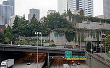 A concrete building with trees and plants hanging from its roof. The building sits on a bridge over a wide freeway, with cars and trucks streaming by.