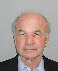 A headshot of an older man facing the camera. The man is wearing a suit without a tie.