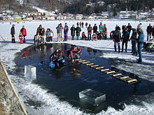 A diver is visible underwater in a hole cut in the ice cover of a small lake. Blocks of ice cut to form the hole are stacked to one side, and a second diver sits on the edge of the hole with his legs in the water. A rough wooden ladder bridges the hole. The dive site is cordoned off with a red and white tape, and other members of the support team stand to the side, with onlookers outside the cordon.