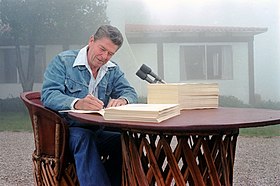 Ronald Reagan sits at a round lattice table with two huge stacks of paper and two microphones. He is wearing denim pants and a denim jacket. He is intently signing one of the stacks of paper. The table is in the gravel driveway in front of a modest timber and plaster house with a tile roof. The house is slightly obscured in fog.