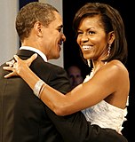 Barack and Michelle Obama are dancing arm-in-arm and smiling. She is wearing a white dress, a large ring, long earrings, and a bracelet. He is wearing a black tuxedo.