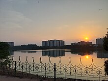 A lake surrounded by a path and fence with apartments. The sun is setting.