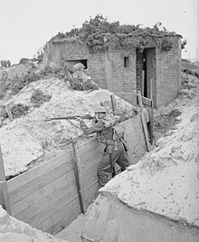 A lone soldier stands in a trench, with his rifle readied on the parapet.