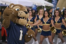 A mascot in a cartoon panther costume wearing an FIU jersey and pointing one finger skyward. Behind him, and blurred in the photo, are cheerleaders in navy blue outfits.