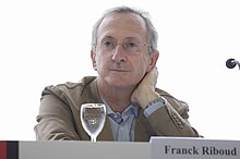 Photographie montrant Franck Riboud, cheveux gris et portant une paire de lunettes discrète, qui se tinet assis, sur la table devant lui, un verre vide et un écriteau où l'on distingue son nom
