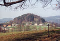 Frontale Farbfotografie von einem Hügel, der mit Bäumen und einem Felsen bestückt ist. Am Rand stehen Häuser und im Vordergrund sind Wiesen mit weiteren Bäumen sowie ein Drahtzaun zu sehen. Hinter dem Hügel sind weitere Hügel zu erkennen.