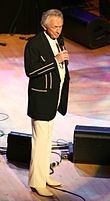 Man in white slacks and black jacket standing singing on stage