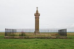 Front-view of a fenced memorial pillar located at Burgh Marsh, topped with a cross. The memorial is said to mark the exact spot where King Edward died.