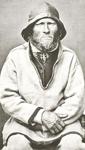Černobílá fotografie sedícího dospělého muže s vousy, s kloboukem a rukama v klíně