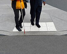 Two people stand on top of light-colored truncated domes at a curb cut. They both face the parking lot, and the person in front holds a white cane.
