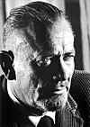 A photo of John Steinbeck. His hair is slicked-back and closely shaved on the sides. He has a mustache and facial hair on his chin.
