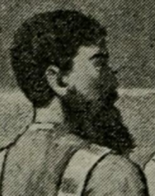The profile of a man, facing right, with a very bushy beard and dark, close-cut hair.