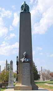 A large obelisk in a graveyard, with a bust of Tyler, and a black cast iron cage partially visible behind it.