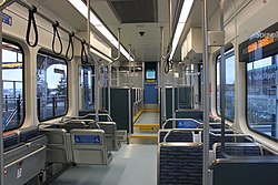 Inside an empty train with large windows, several seats arranged parallel and perpendicularly, metal handlebars, and black hand-holds. The far section of the train with additional seating is lifted up and has a short stairway.