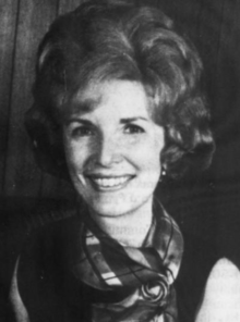 A smiling white woman with her hair in a bouffant style, wearing a print scarf and a dark sleeveless dress