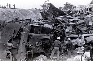 Workers standing around a destroyed freight locomotive, destroyed hopper cars are piled behind the locomotive. Several workers overlook the scene from the railroad track embankment.
