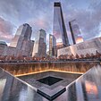 Illuminated water falls into the square 9/11 Memorial south pool at sunset, and glass-clad One World Trade Center and other skyscrapers rise in the background