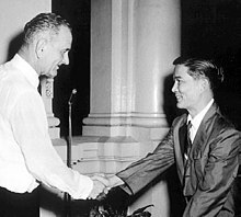 Tall European man standing in profile at left in a white suit and tie shakes hands with a smaller black-haired Asian man in a white shirt, dark suit and tie.