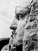 Profile of stone face jutting out from a mountainside. Three workers clamber over it, each about the height of the face's upper lip.